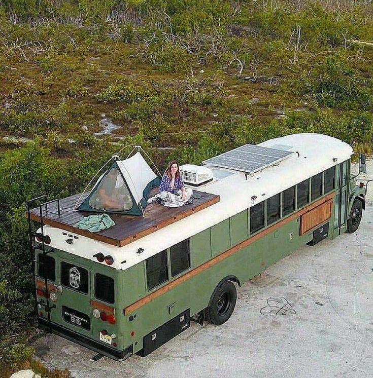 a woman sitting on top of a green bus next to a forest filled with trees