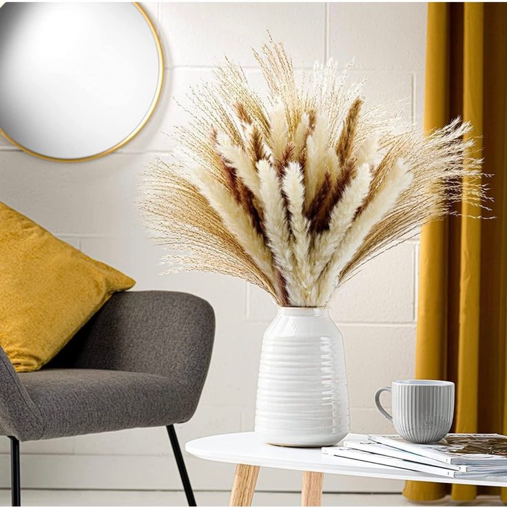 a white vase filled with dry grass sitting on top of a table next to a gray chair