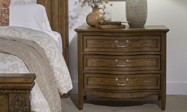 a bedroom scene with focus on the nightstand and bed in the foreground, there is a vase sitting on top of the dresser