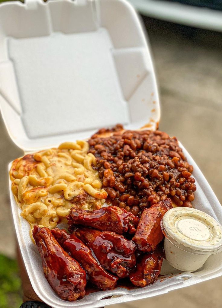 a person holding a takeout box filled with meat, beans and macaroni