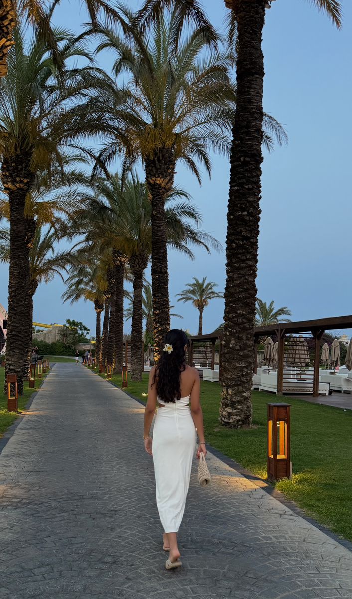 a woman in a white dress walking down a path between palm trees