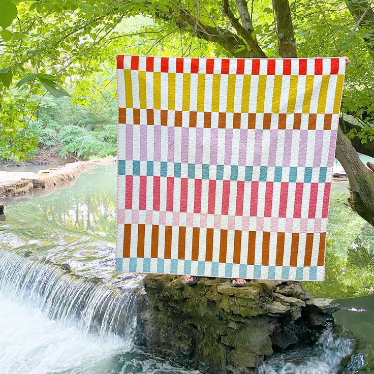 a colorful quilt hanging over a waterfall in the woods