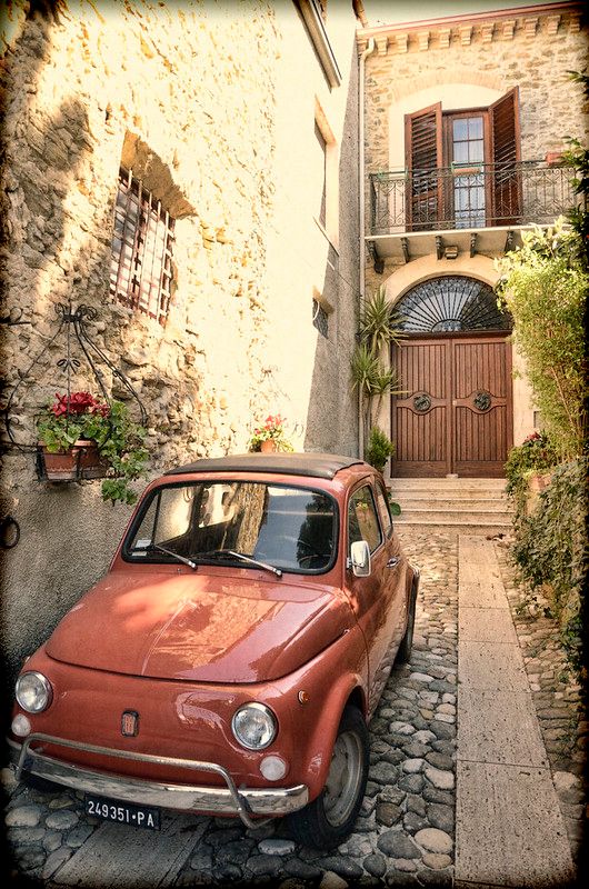 an old red car parked in front of a stone building with shutters and windows