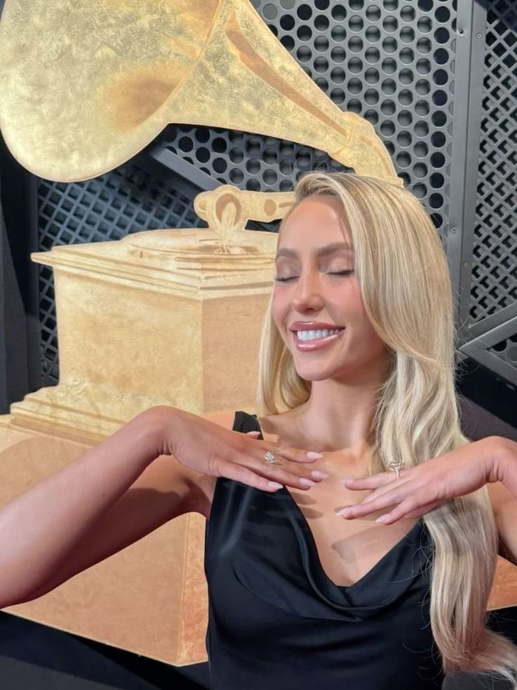 a woman with long blonde hair standing in front of a large golden trophy and wearing a black dress