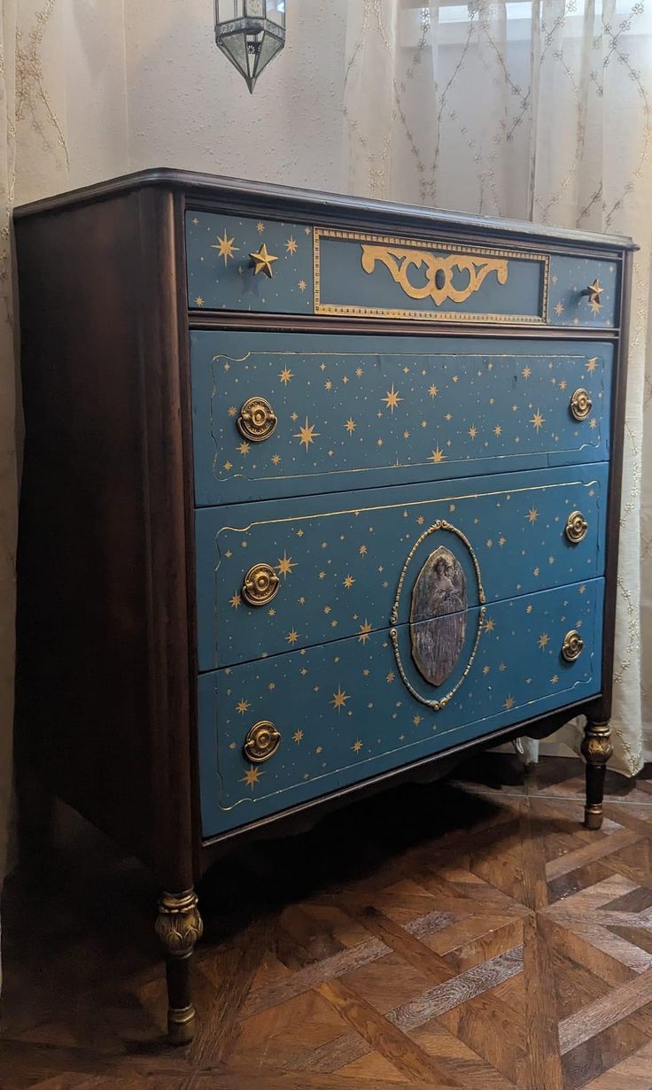 a blue dresser with gold stars painted on the top and bottom, sitting in front of a window