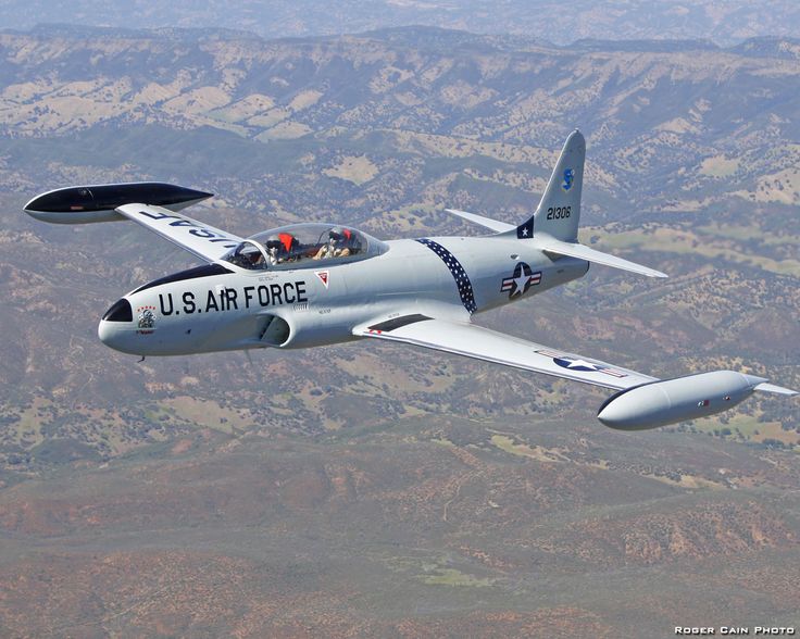 a u s air force plane flying in the sky with mountains and trees behind it