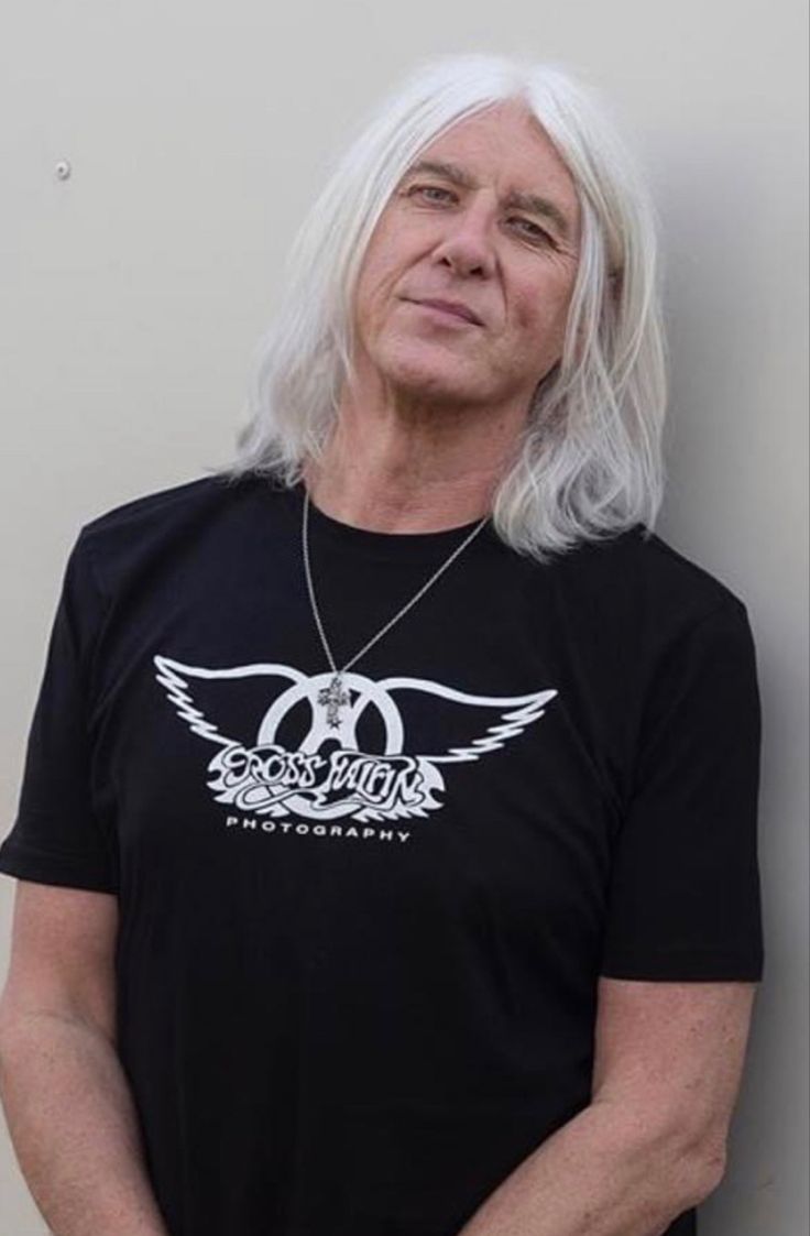 an older man with white hair wearing a black t - shirt and standing against a wall