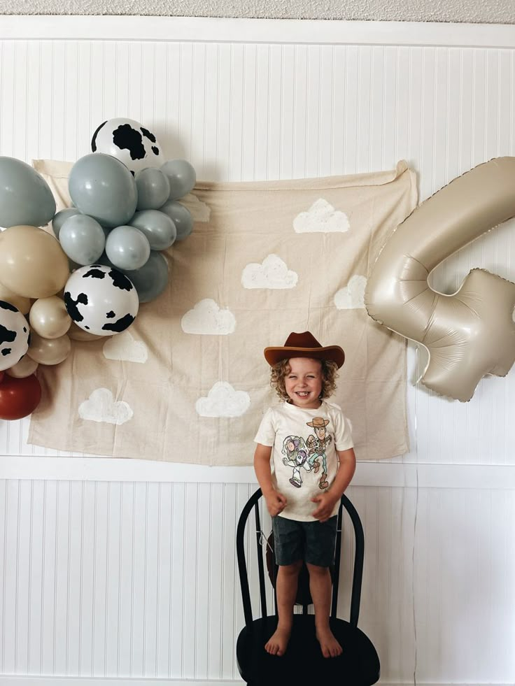 a little boy wearing a cowboy hat standing on a chair in front of a wall with balloons