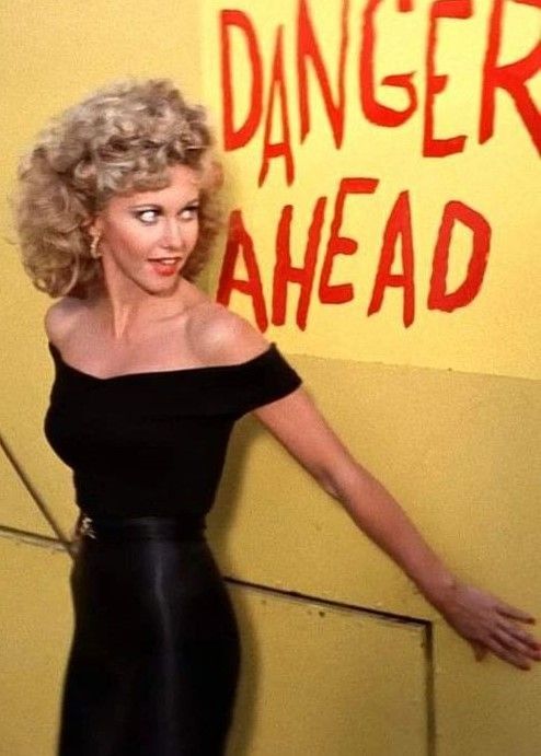 a woman with blonde hair and black dress posing in front of a sign that says danger ahead