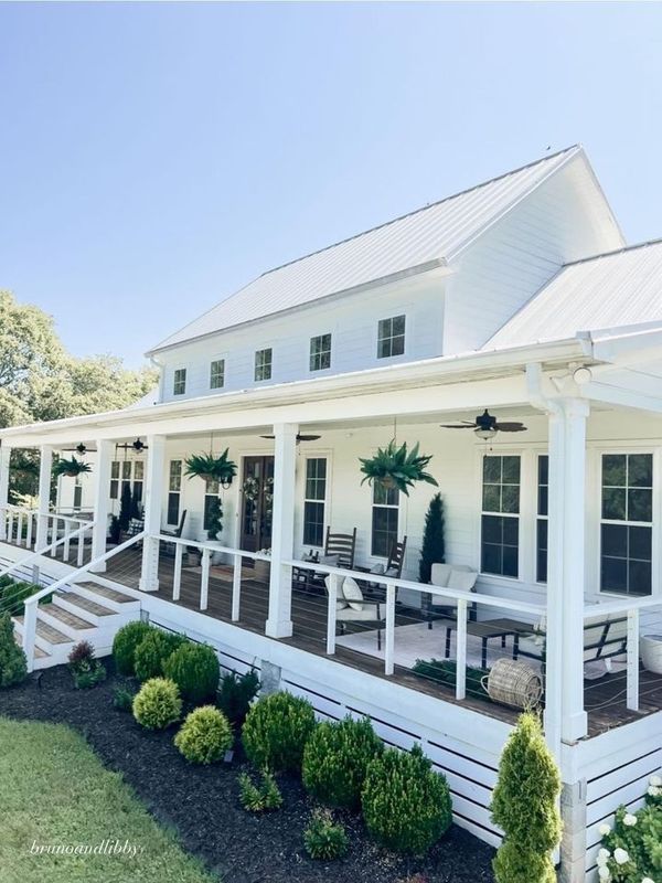 a white house with porches and chairs on the front lawn, surrounded by shrubs