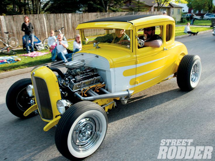 an old yellow car driving down the street with people in the back ground looking at it