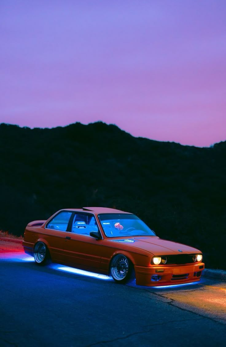 an orange car is parked on the side of the road with its lights on at night