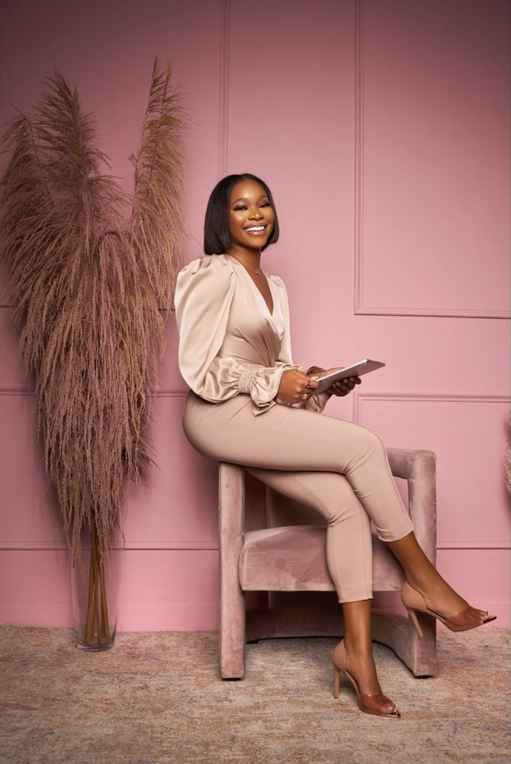 a woman sitting on a chair in front of a pink wall and holding a tablet