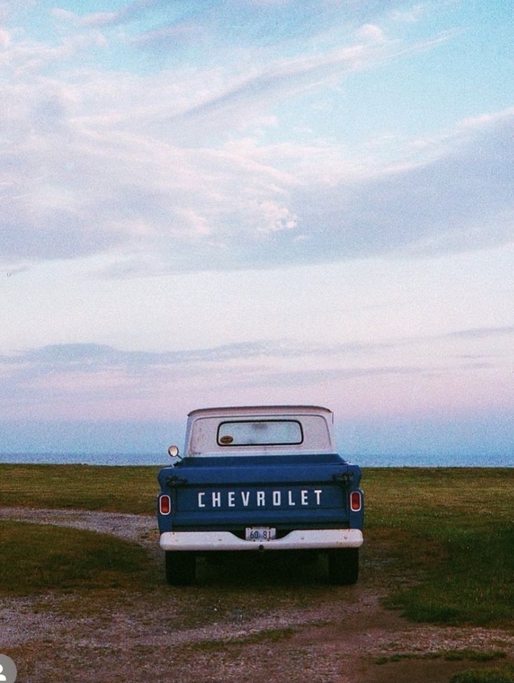 an old chevy truck parked on the side of a dirt road next to a grassy field