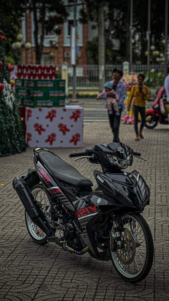 a black motorcycle parked on the side of a brick road with people walking around it