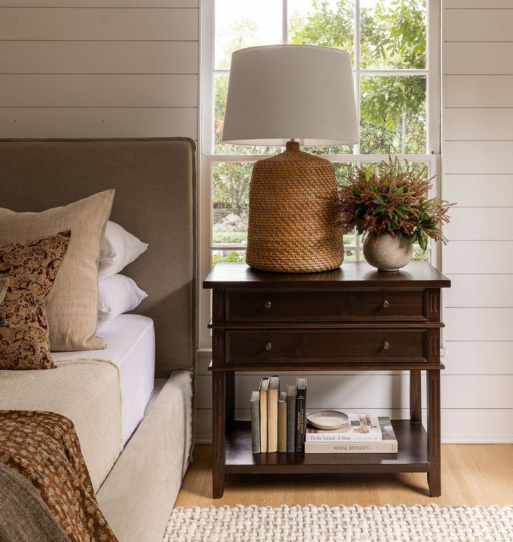 a bedroom with a bed, nightstand and lamp on it's side table in front of a window