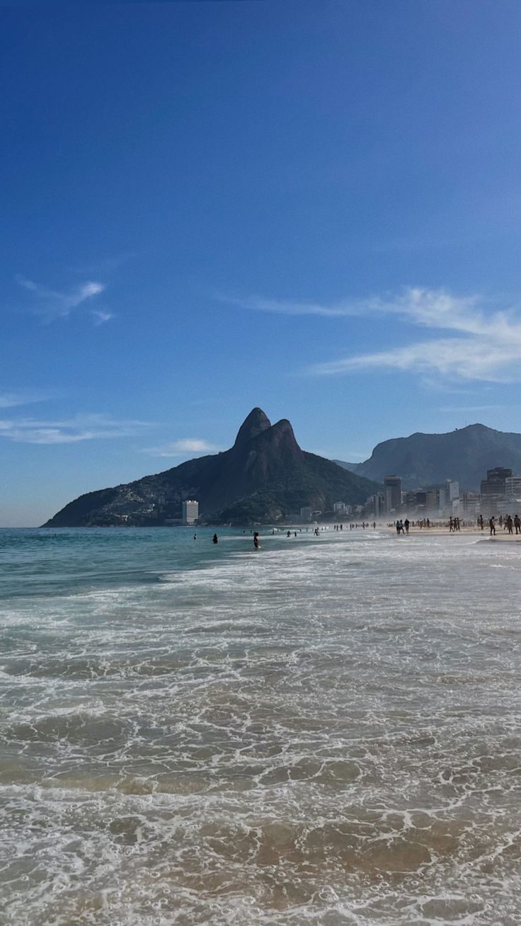 some people are walking on the beach and in the water with mountains in the background