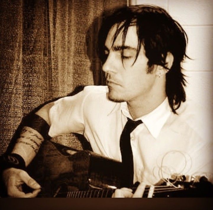 a man with long hair wearing a white shirt and black tie playing an acoustic guitar