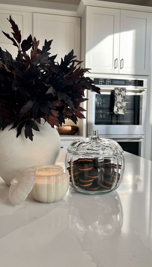 a white vase filled with leaves next to a candle on top of a kitchen counter