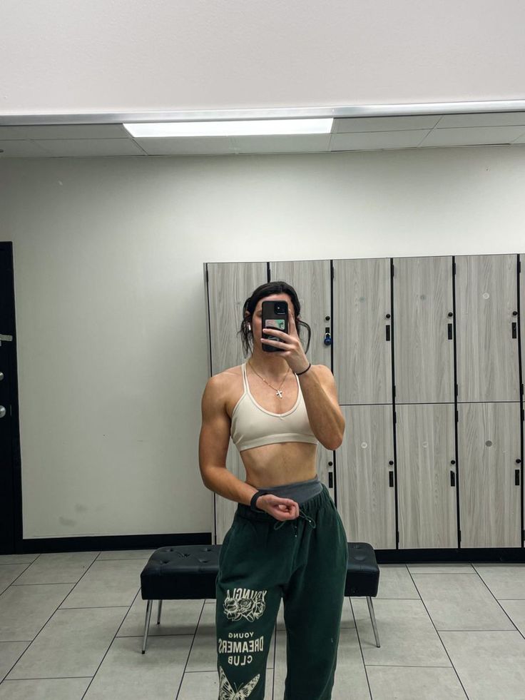 a woman taking a selfie in front of lockers wearing green sweatpants and a white tank top