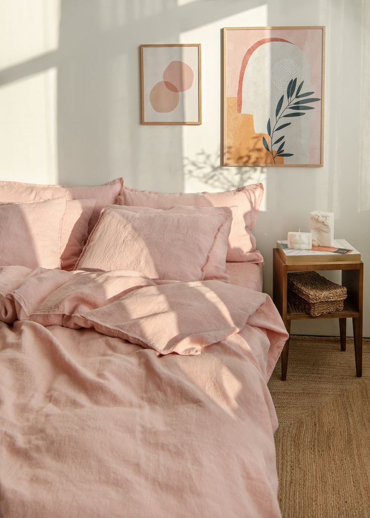 a bed with pink linens and pillows in a white room next to a wooden table