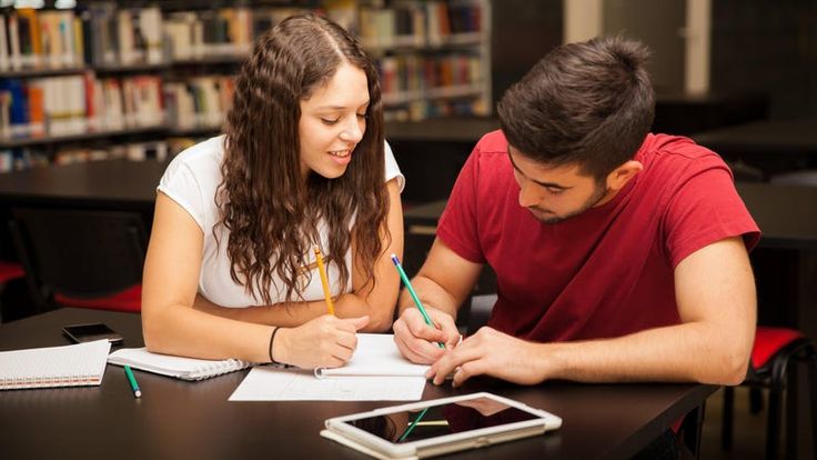 a man and woman sitting at a table in front of a tablet computer writing on paper