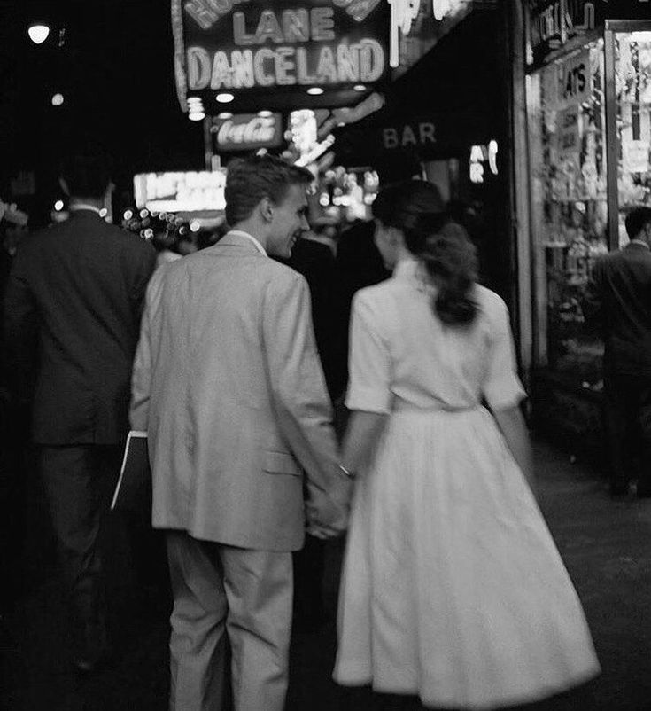 a man and woman walking down a street holding hands in front of a neon sign