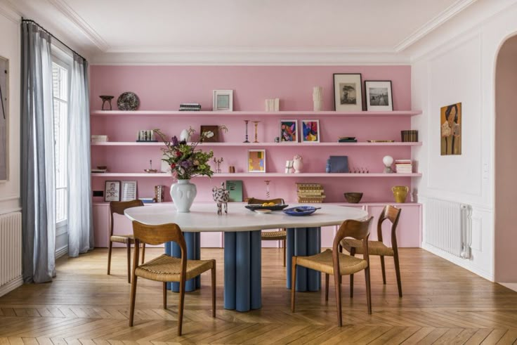 a dining room with pink walls and wooden chairs in front of a white table surrounded by bookshelves