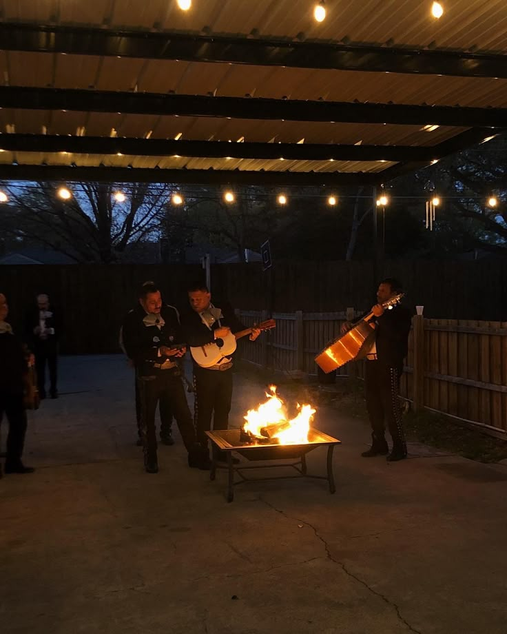 some people are playing guitar and singing around a fire pit with lights hanging from the ceiling