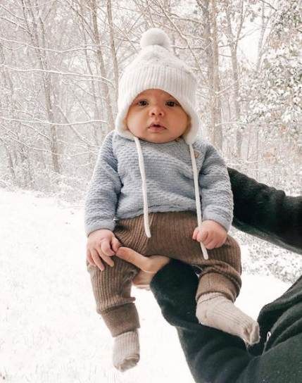 a man holding a baby wearing a hat and sweater in the snow with trees behind him