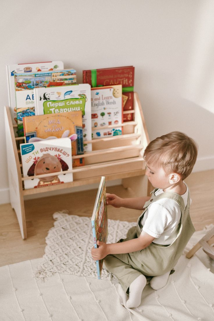 a young boy sitting on the floor reading a book in front of a wooden shelf