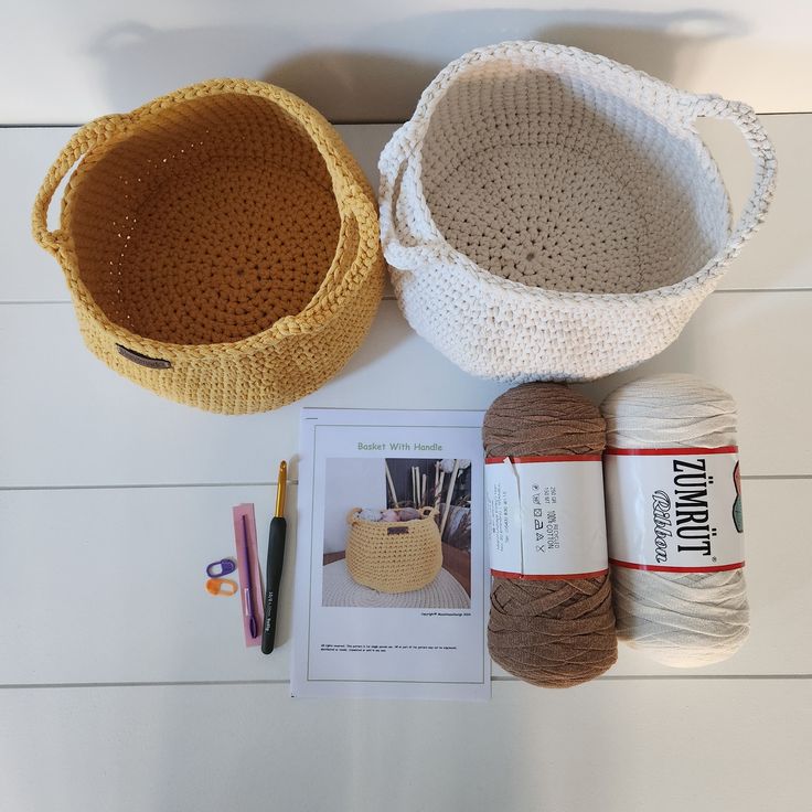 two crocheted baskets, yarn and scissors on a white wooden table with an instruction book