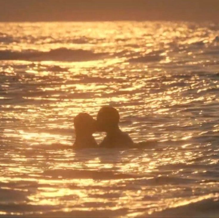 two people swimming in the ocean at sunset with their backs to each other and kissing