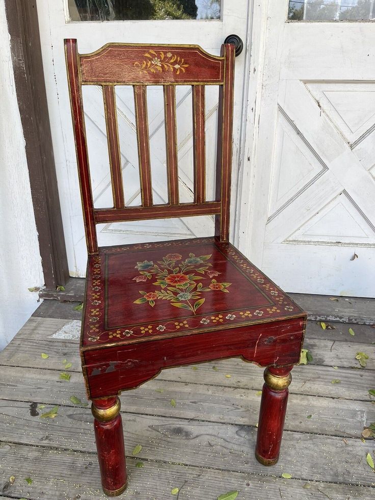 an old wooden chair painted red and gold with flowers on the seat is sitting in front of a white door