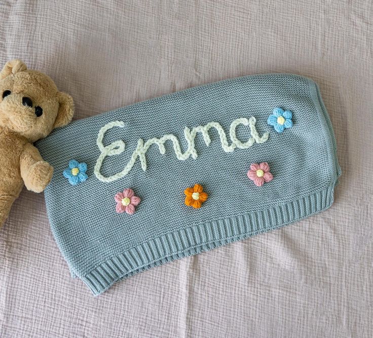 a teddy bear laying on top of a bed next to a knitted name sign