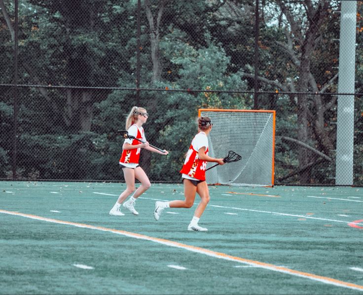 two girls in red and white uniforms are playing lacrosse on the field with trees in the background