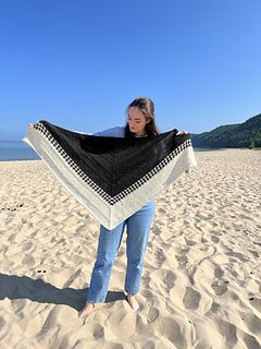 a woman standing on top of a sandy beach holding a black and white shawl