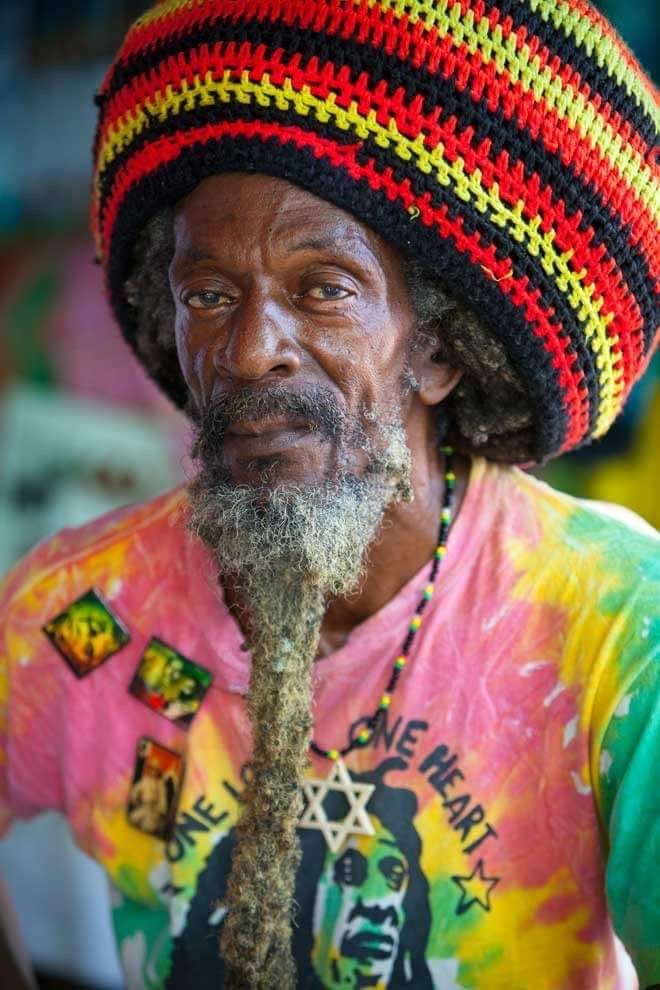 a man with a beard wearing a colorful hat and tie dye t - shirt looks at the camera