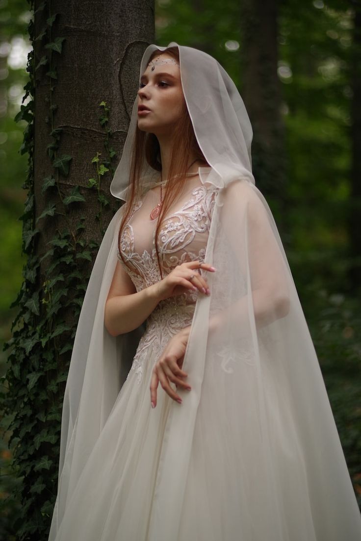 a woman in a white wedding dress and veil standing next to a tree with ivy growing on it