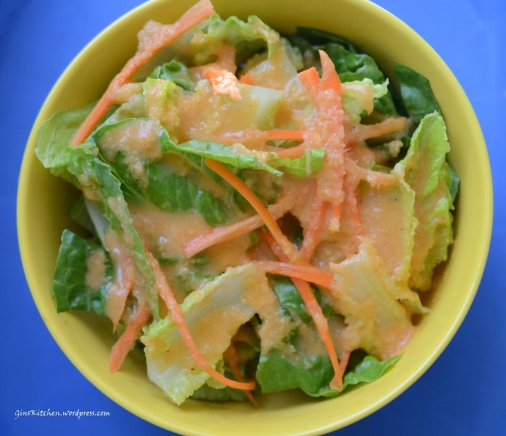 a yellow bowl filled with lettuce, carrots and dressing on top of a blue table