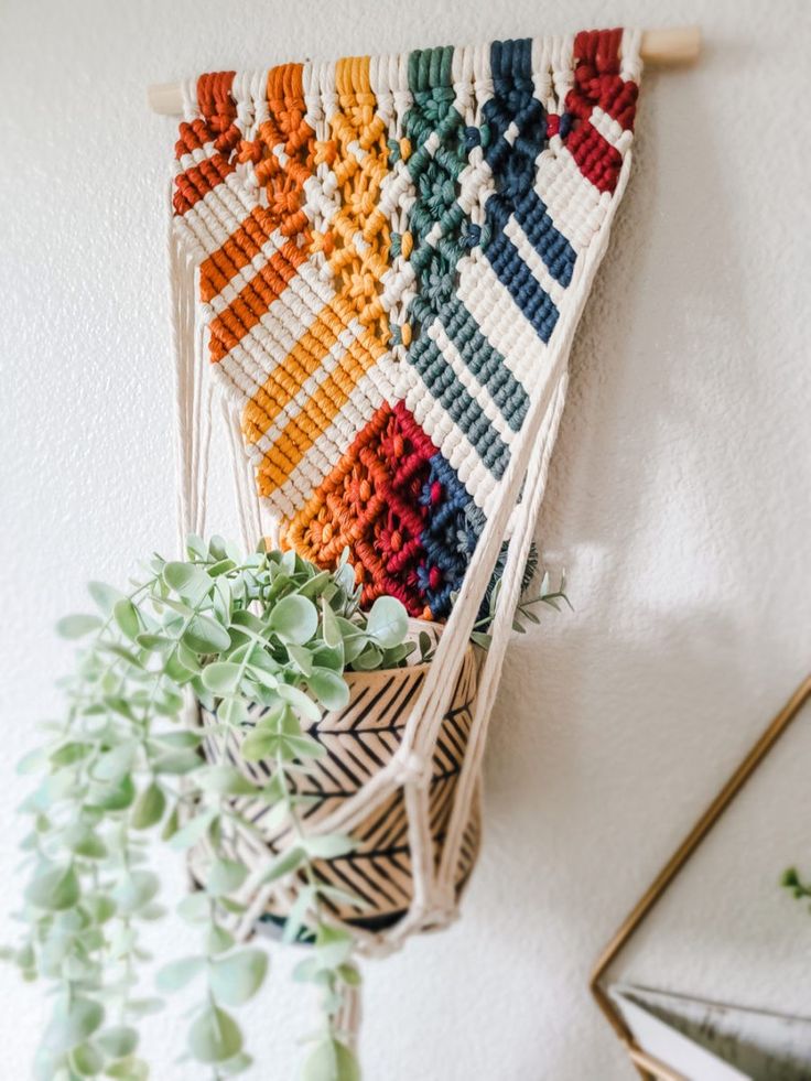 a wall hanging made out of different colors of yarn and wood with plants in the corner