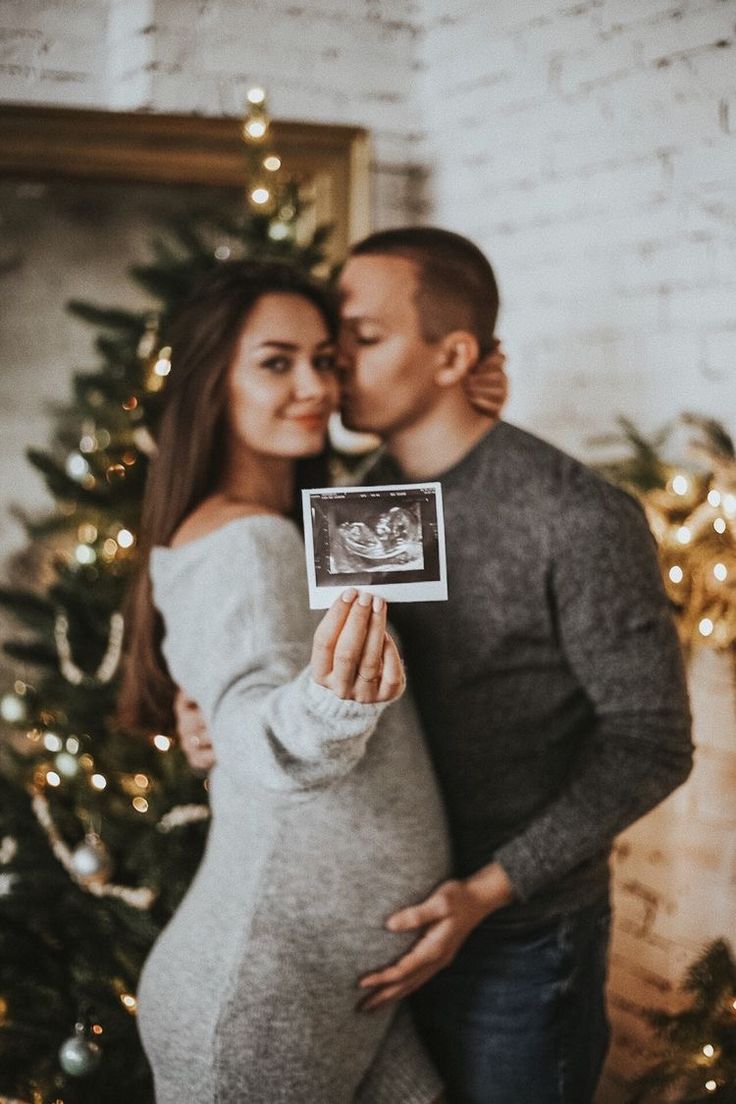 a pregnant couple standing in front of a christmas tree and holding up an old photo