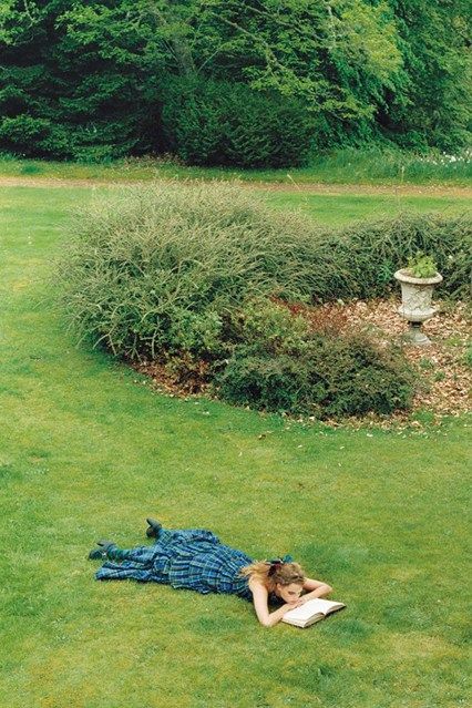 a woman laying on the ground reading a book in a field with trees and bushes behind her