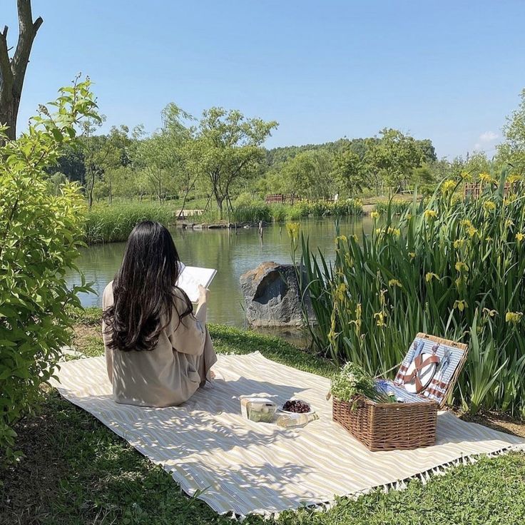 a woman sitting on a blanket in front of a pond reading a book and painting