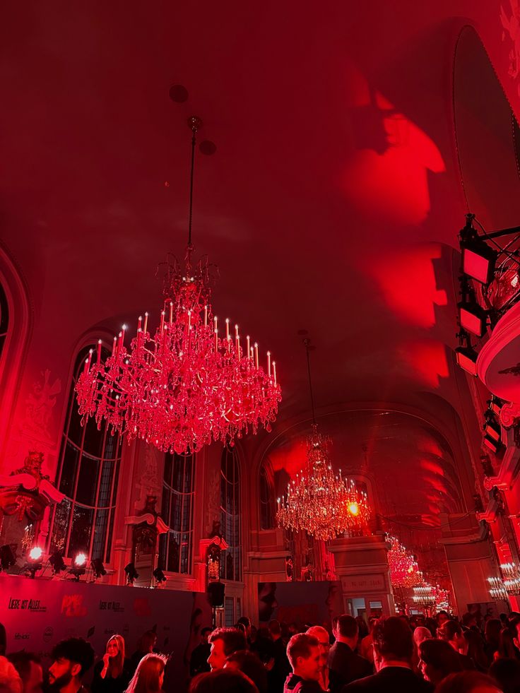 a large chandelier hanging from the ceiling in a room with red lighting and people standing around