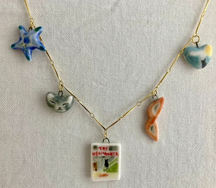 Vintage-themed charm necklace on a gold bar chain; all the charms are sculpted from a fine white porcelain clay and painted by hand to have charming details. Back and front sides are painted. Since they are made from porcelain and glossed with a clear glaze, each charm is smooth and soft to the touch.  Each charm is attached to a small, gold jump ring and can be attached to a necklace chain. Small charms featured: New Yorker cover with cat looking out at city, Curled up cat sleeping, Green Star Fish, Blue Star Fish, Love Letter w/ gold highlights, Red Lipstick with gold band, Red lips, Slipper with gold heart, Gold heart (gold raindrops on one side, gold heart on the other), Lucky Strikes pack, Moon and Black Cat on a heart, Sun Hat with gold band. Bought individually, your charm(s) will c Clay Star Necklace, Homemade Charms, Diy Initial Necklace, Curled Up Cat, Clay Charm Necklace, Clay Chain, Highlights Red, New Yorker Cover, Lucky Charm Necklace