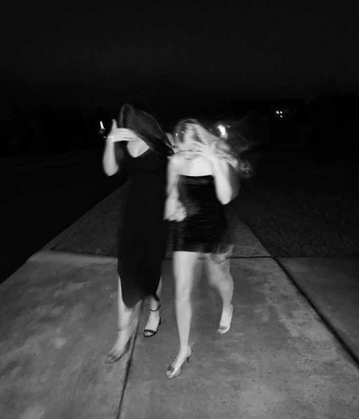 two young women are dancing on the sidewalk at night, one is wearing a black dress