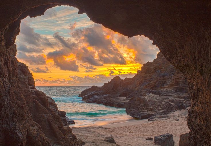 the sun is setting over the ocean from inside a cave on the beach with rocks and sand