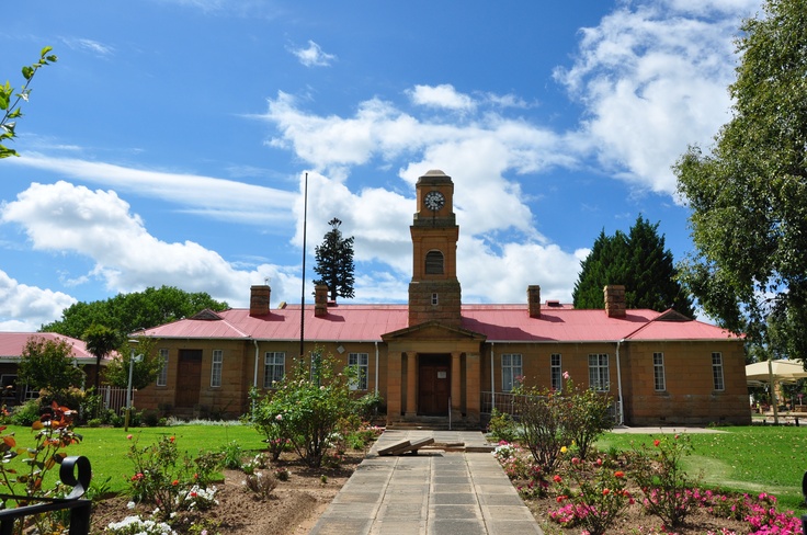 an old brick building with a clock tower in the center and flowers around it on a sunny day