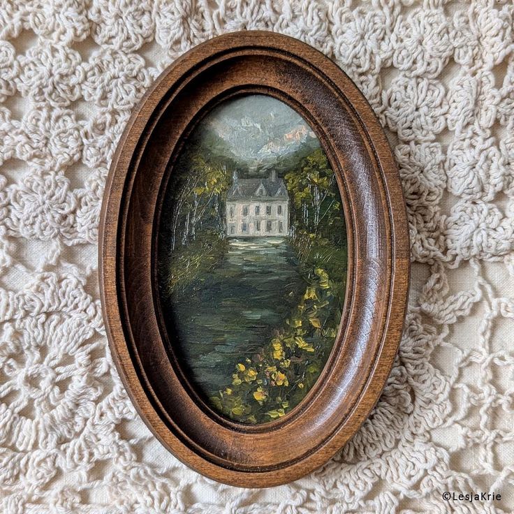 an oval wooden frame with a painting of a house in the background on a lace covered surface